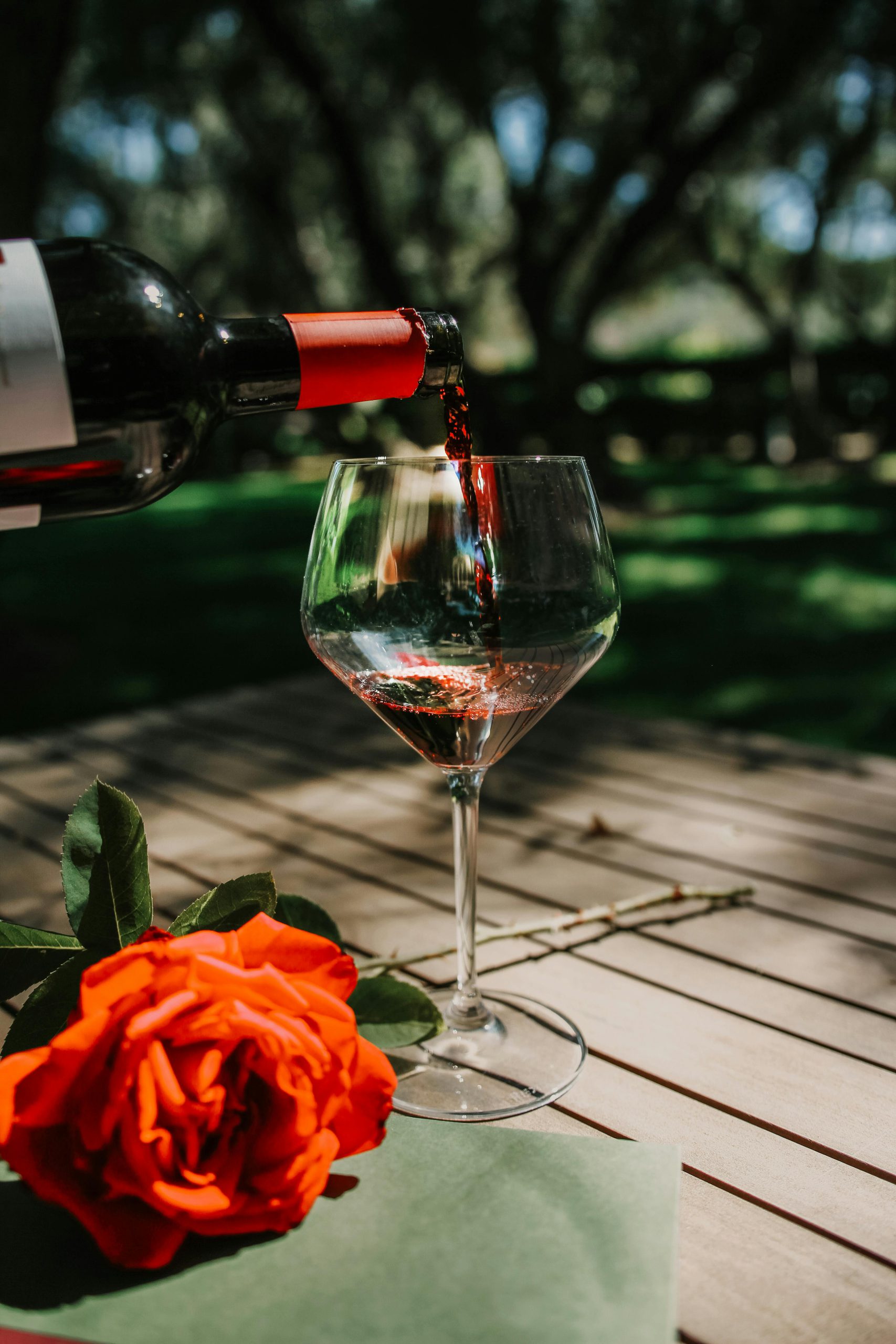 Vibrant red wine being poured into a glass on a table with a red rose, set outdoors in Napa.