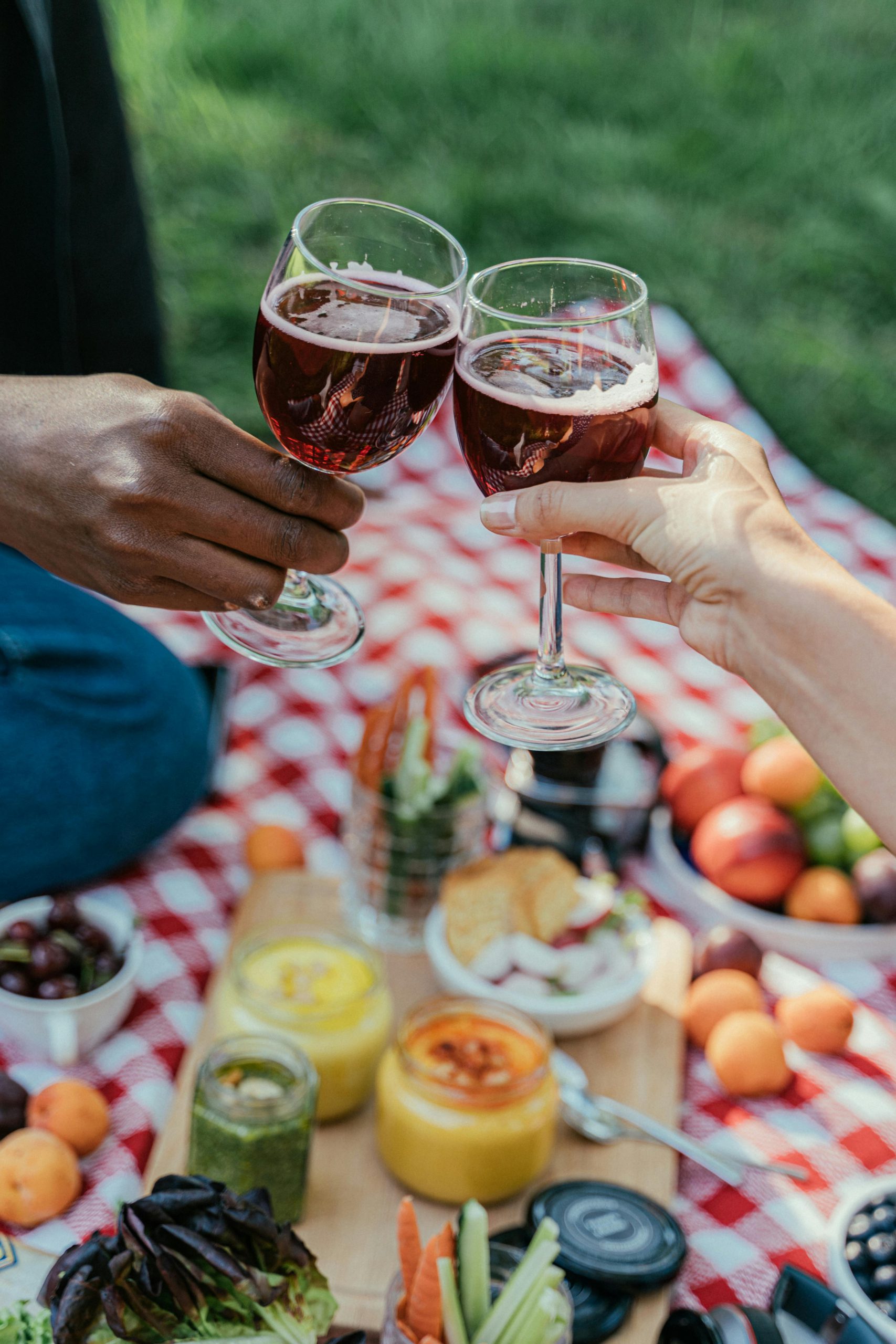 Two people clinking wine glasses over a vibrant picnic setting outdoors.